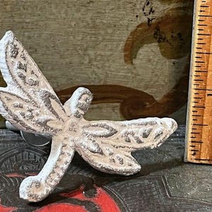 May include: A decorative dragonfly-shaped cabinet knob with a distressed white and brown finish. The knob is made of metal and features detailed wing and body patterns. A wooden ruler is next to the knob.