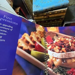 May include: A cookbook titled "First Impressions" with a blue cover and the text "An appealing assortment of awesome, attention-getting appetizers". The open book shows a recipe for a baked brie with cranberries, almonds, and rosemary.