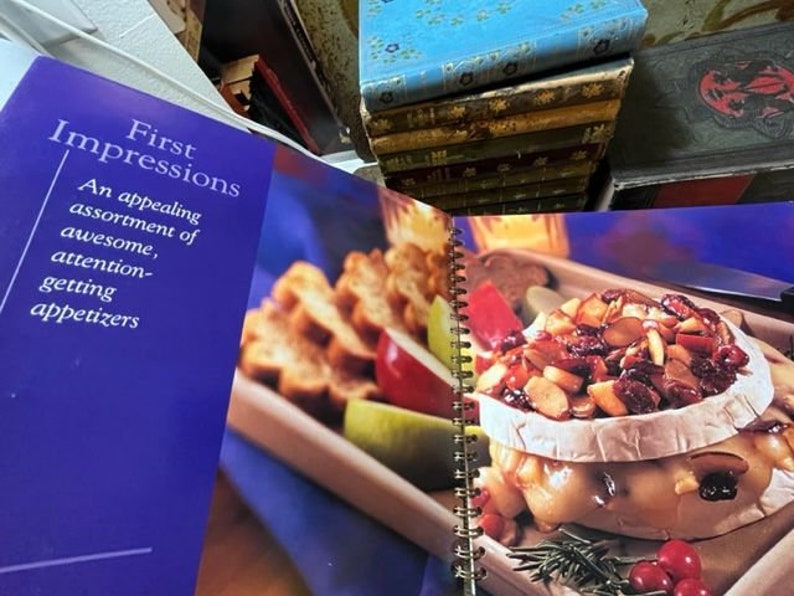 May include: A cookbook titled "First Impressions" with a blue cover and the text "An appealing assortment of awesome, attention-getting appetizers". The open book shows a recipe for a baked brie with cranberries, almonds, and rosemary.