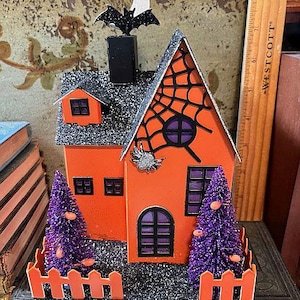 May include: A Halloween-themed decorative house with an orange facade, black roof, and a spiderweb design. Features a bat and moon on top, purple trees, and an orange picket fence. The house has black windows and a door.
