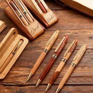 May include: Wooden pens with gold accents and personalized names are displayed on a wooden surface. The pens are in wooden cases, some with two pens and others with one. The names "David", "Rachel", "David Bell", "Lana Said", "Amelie Wedrich", and "Julian Bondel" are engraved.
