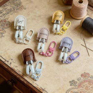 Könnte beinhalten: Eine Sammlung personalisierter Schlüsselanhänger in verschiedenen Farben, darunter Braun, Beige, Gelb, Rosa und Lila. Jeder Schlüsselanhänger hat ein Lederetikett mit einem Namen und einen Metallverschluss. Die Schlüsselanhänger sind auf einer gemusterten Oberfläche angeordnet.