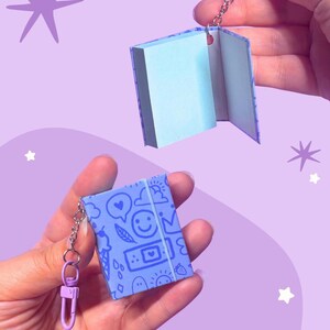 May include: A miniature book keyring featuring a light purple cover with a repeating pattern of blue line art icons including a smiley face, ice cream, clouds, and a bandage. The keyring includes a light purple lobster clasp and metal chain.