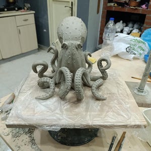 May include: A light grey clay sculpture of an octopus with a textured mantle and multiple tentacles featuring visible suction cups. The sculpture is positioned on a square wooden board covered with a thin, clear plastic sheet for protection.