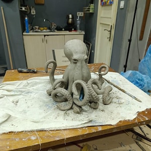 Op de afbeelding: Een grijze klei octopus sculptuur met gedetailleerde tentakels, rustend op een met witte doek bedekte tafel. De sculptuur is het middelpunt en toont de vaardigheid van de kunstenaar. De achtergrond omvat een werkruimte met gereedschap en kunstbenodigdheden.