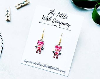 May include: A pair of pink robot earrings with gold accents and heart-shaped eyes. The earrings are on a white card with the text "The Little Wish Company" printed on it.