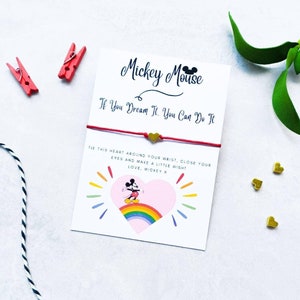 Könnte beinhalten: Ein rotes und weißes Armband aus Schnur mit einem goldenen Herz-Anhänger und einer Mickey-Maus-Figur auf einem Regenbogen. Die Karte lautet "Mickey Mouse Wenn du es träumst, kannst du es tun. Binde dieses Herz um dein Handgelenk, schließe deine Augen und wünsch dir etwas! Liebe, Mickey X."