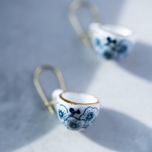 Puede incluir: Un par de pendientes de aro dorados con un dije de taza de té de porcelana blanca. Las tazas de té tienen un patrón floral azul y blanco.