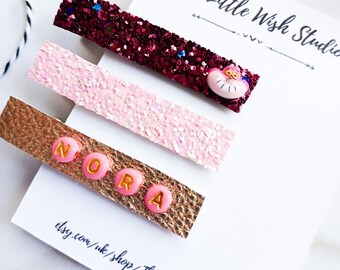 Op de afbeelding: Drie haarclips met glitter en een roze en gouden lederen basis. De eerste clip heeft rode glitter en een roze kattengezicht bedel. De tweede clip heeft roze glitter. De derde clip heeft gouden glitter en de naam "Nora" gespeld in roze letters.