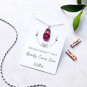 May include: A silver necklace with a small, colorful, painted wooden doll pendant. The necklace is displayed on a white card with the text "Never forget that beauty comes from within."