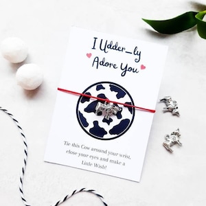 Puede incluir: Un brazalete de cuerda roja con un dije de vaca y una tarjeta blanca con el texto "I Udder-ly Adore You" e instrucciones para atar el brazalete alrededor de tu muñeca, cerrar los ojos y pedir un deseo.