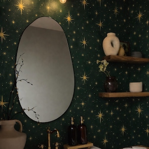 Puede incluir: Interior de baño con pared verde oscuro con estampado de estrellas doradas. Un espejo ovalado cuelga sobre un lavabo blanco con grifo dorado. Dos botellas marrones sobre una bandeja de madera. Una lámpara con dos pantallas doradas está montada encima del espejo.