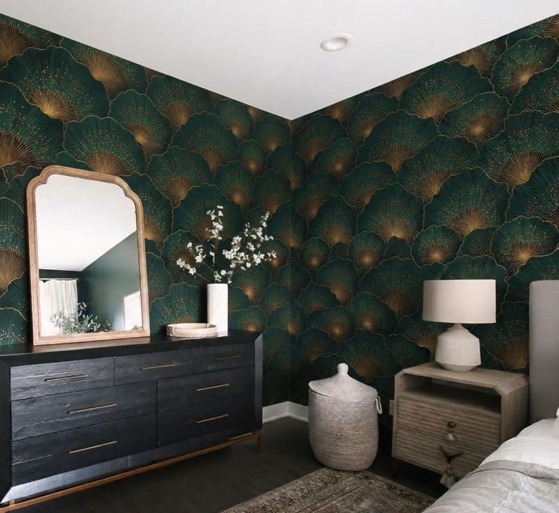 May include: A bedroom with dark green and gold patterned wallpaper. A black dresser with gold hardware and a wooden-framed mirror are against the wall. A white lamp sits on a wooden nightstand next to a bed with a gray headboard.