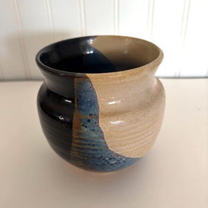 May include: A small ceramic pot with a rounded body and a flared rim. The exterior features a two-tone glaze, with one side in a glossy dark brown and the other in a matte beige, separated by a vertical band of speckled blue and brown glaze.