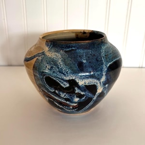 May include: A round ceramic vase featuring a glossy glaze with a blend of deep navy blue, black, and white swirling patterns, contrasted by a smooth tan section on the left side. The vessel has a wide, open mouth and a rounded, bulbous body shape.