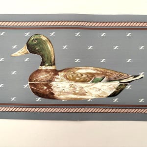 May include: A wallpaper border featuring a mallard duck with a green head and brown body centered on a dusty blue background. The background is patterned with small white symbols and framed by horizontal bands with a rope-like texture in tan.