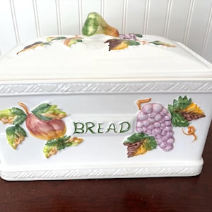 May include: A rectangular white ceramic bread box with a removable lid featuring a pear-shaped handle. The front displays a painted design of a peach and a cluster of purple grapes with green leaves, with the word BREAD centered in green text.