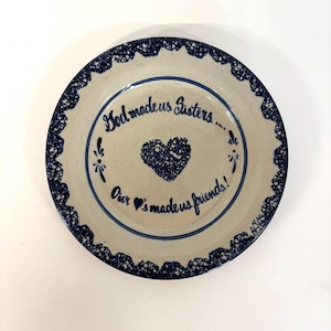 Op de afbeelding: Een rond keramieken bord met een gespikkelde beige basis en een donkerblauwe kantachtige rand. Het midden bevat een donkerblauw hart en de tekst: God made us Sisters.... Our hearts made us friends!