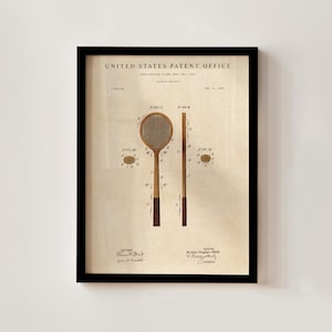 Op de afbeelding: Ingelijste vintage tennisracket patent print. De print toont een gedetailleerde illustratie van een tennisracket en zijn onderdelen, met de woorden "United States Patent Office" bovenaan. De print is in sepia tinten en ingelijst in een zwart frame.