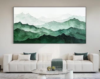 May include: A large, rectangular abstract painting of layered green mountains against a white sky. The artwork features varying shades of green, creating depth and texture. The painting is framed in black and hangs above a white sofa.