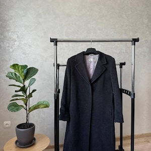 May include: A long, dark gray coat hanging on a black metal clothing rack. The coat has a classic design with a collar, lapels, and pockets.