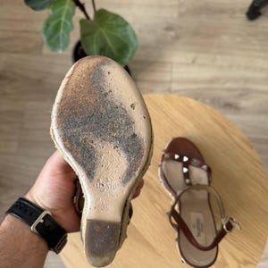 May include: A pair of brown leather wedge sandals with metallic studs and an espadrille-style sole. The sole of one sandal is visible, showing the Valentino brand name, the size 36, and the text Made in Italy embossed on the beige base.
