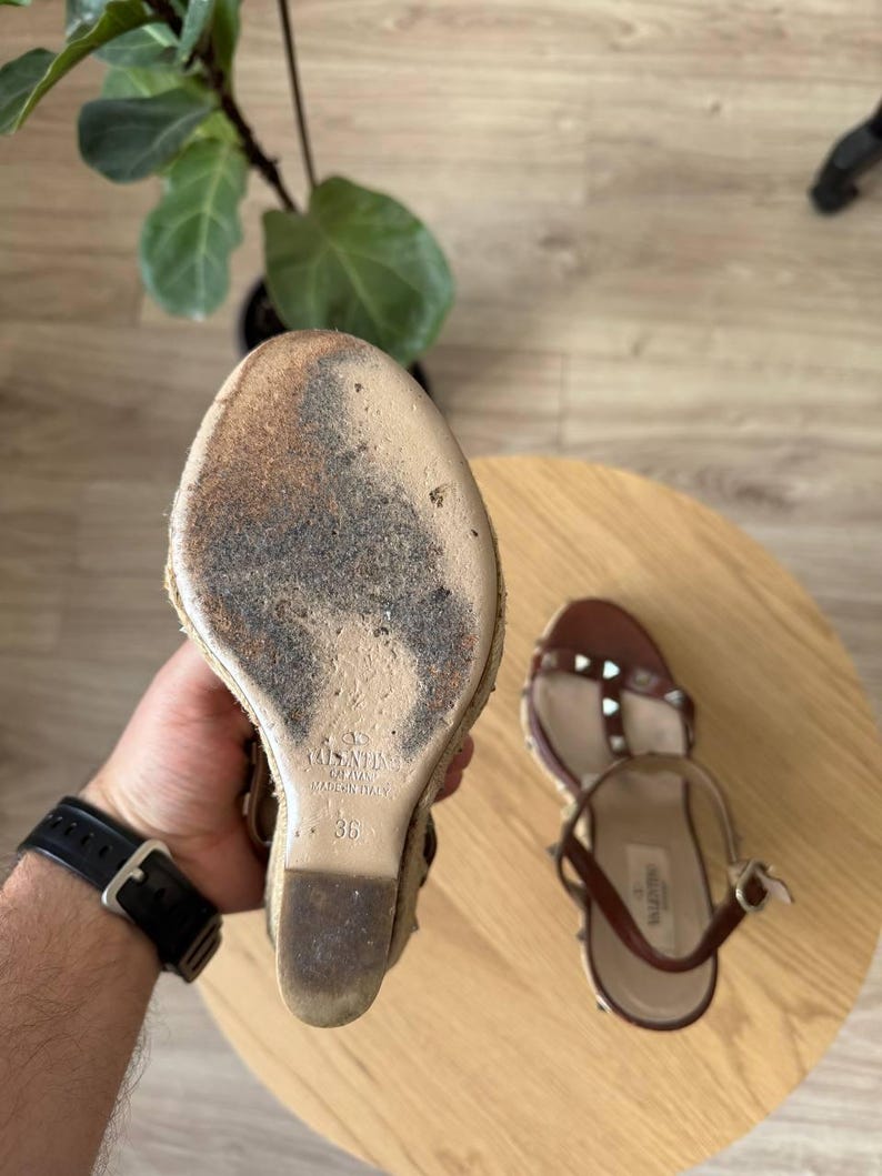 May include: A pair of brown leather wedge sandals with metallic studs and an espadrille-style sole. The sole of one sandal is visible, showing the Valentino brand name, the size 36, and the text Made in Italy embossed on the beige base.
