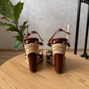 May include: A pair of brown leather wedge sandals featuring a jute-wrapped heel and platform. The sandals are adorned with silver-tone pyramid studs along the straps and the base of the heel, and include an adjustable ankle strap with a metal buckle.