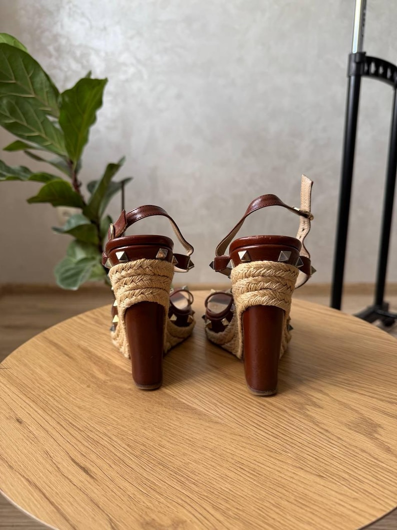 May include: A pair of brown leather wedge sandals featuring a jute-wrapped heel and platform. The sandals are adorned with silver-tone pyramid studs along the straps and the base of the heel, and include an adjustable ankle strap with a metal buckle.