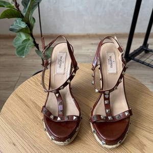 May include: A pair of brown leather wedge sandals featuring a t-strap design, silver-tone pyramid studs, and an adjustable ankle strap with a buckle. The insoles are labeled with the Valentino Garavani brand name, and the soles are wrapped in jute.