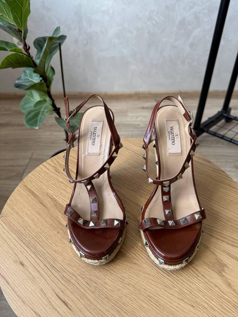 May include: A pair of brown leather wedge sandals featuring a t-strap design, silver-tone pyramid studs, and an adjustable ankle strap with a buckle. The insoles are labeled with the Valentino Garavani brand name, and the soles are wrapped in jute.