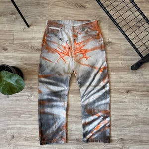 Może przedstawiać: Para jasnych dżinsów z pomarańczowo-szarym wzorem tie-dye. Dżinsy mają pomarańczowy wzór rozbłysku na przodzie i spiralny wzór na nogawkach. Unikalny, artystyczny styl.