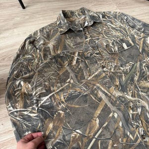 May include: Camouflage button-down shirt in shades of brown, green, and tan. The long-sleeved shirt features two chest pockets and a collared neckline. The fabric has a detailed pattern designed to blend with outdoor environments.
