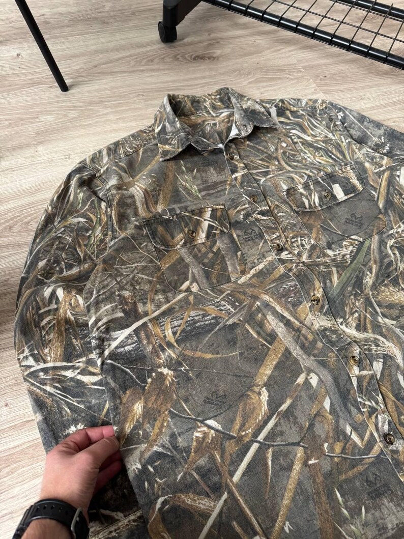 May include: Camouflage button-down shirt in shades of brown, green, and tan. The long-sleeved shirt features two chest pockets and a collared neckline. The fabric has a detailed pattern designed to blend with outdoor environments.