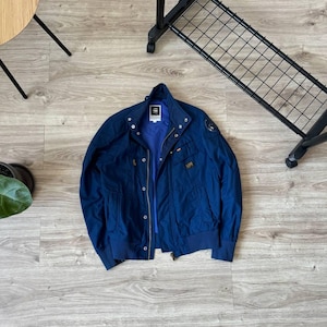 May include: A dark blue bomber jacket with a zipper closure and multiple pockets. The jacket has a G-Star Raw logo on the left sleeve.