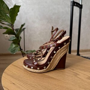 May include: A pair of brown leather wedge sandals featuring a jute-wrapped platform sole and gold-tone pyramid studs along the straps. The sandals have an adjustable ankle strap with a buckle closure and a high wedge heel.
