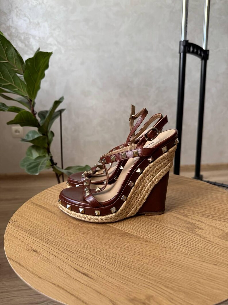 May include: A pair of brown leather wedge sandals featuring a jute-wrapped platform sole and gold-tone pyramid studs along the straps. The sandals have an adjustable ankle strap with a buckle closure and a high wedge heel.