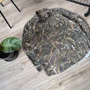 May include: A long-sleeved button-down shirt with a camouflage pattern in shades of brown, green, and tan. The shirt has two chest pockets and a collared neckline. The fabric appears to be a lightweight material.
