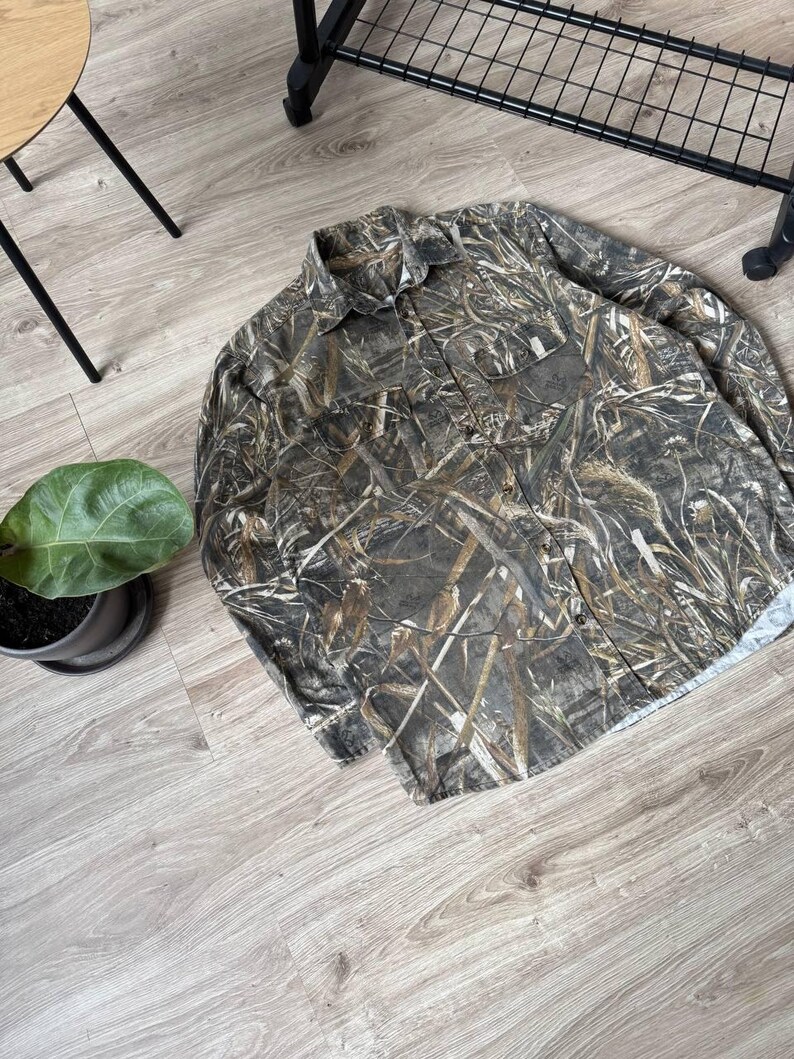 May include: A long-sleeved button-down shirt with a camouflage pattern in shades of brown, green, and tan. The shirt has two chest pockets and a collared neckline. The fabric appears to be a lightweight material.
