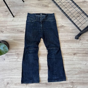 May include: Dark wash denim jeans with a high waist and flared leg. The jeans have a button and zip fastening and feature unique seam details on the knees. The jeans are laid flat on a wooden floor.