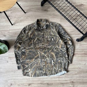May include: A long-sleeved button-down shirt with a camouflage pattern in shades of brown, green, and tan. The shirt has two chest pockets and a classic collar. The fabric appears to be a medium-weight material.