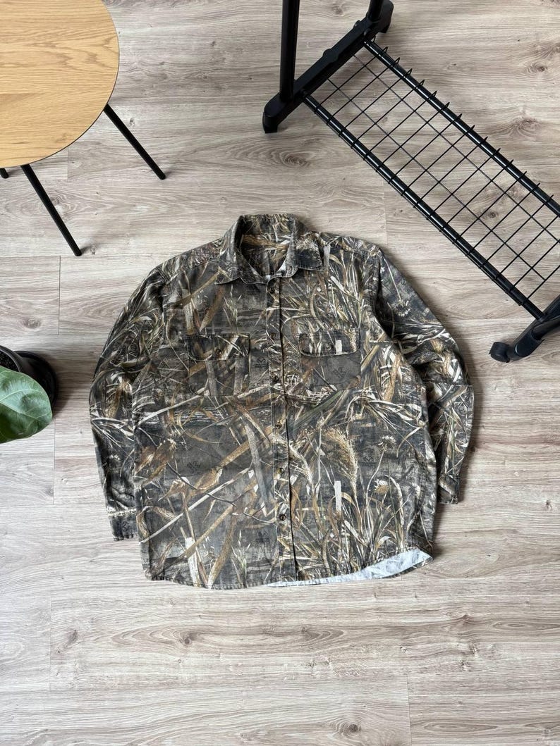 May include: A long-sleeved button-down shirt with a camouflage pattern in shades of brown, green, and tan. The shirt has two chest pockets and a classic collar. The fabric appears to be a medium-weight material.