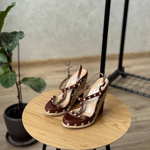 May include: A pair of brown leather wedge sandals featuring a slingback strap, an open toe, and a jute-wrapped wedge heel. The straps are adorned with small, metallic pyramid-shaped studs, and the insole is branded with the Valentino logo.
