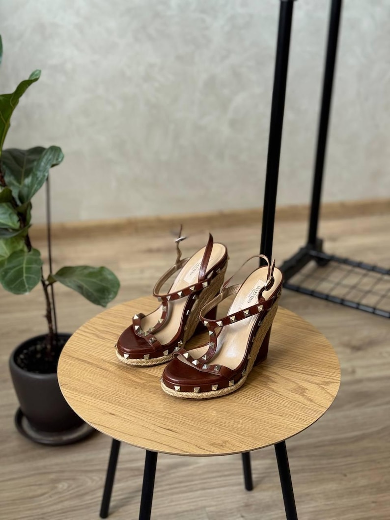 May include: A pair of brown leather wedge sandals featuring a slingback strap, an open toe, and a jute-wrapped wedge heel. The straps are adorned with small, metallic pyramid-shaped studs, and the insole is branded with the Valentino logo.