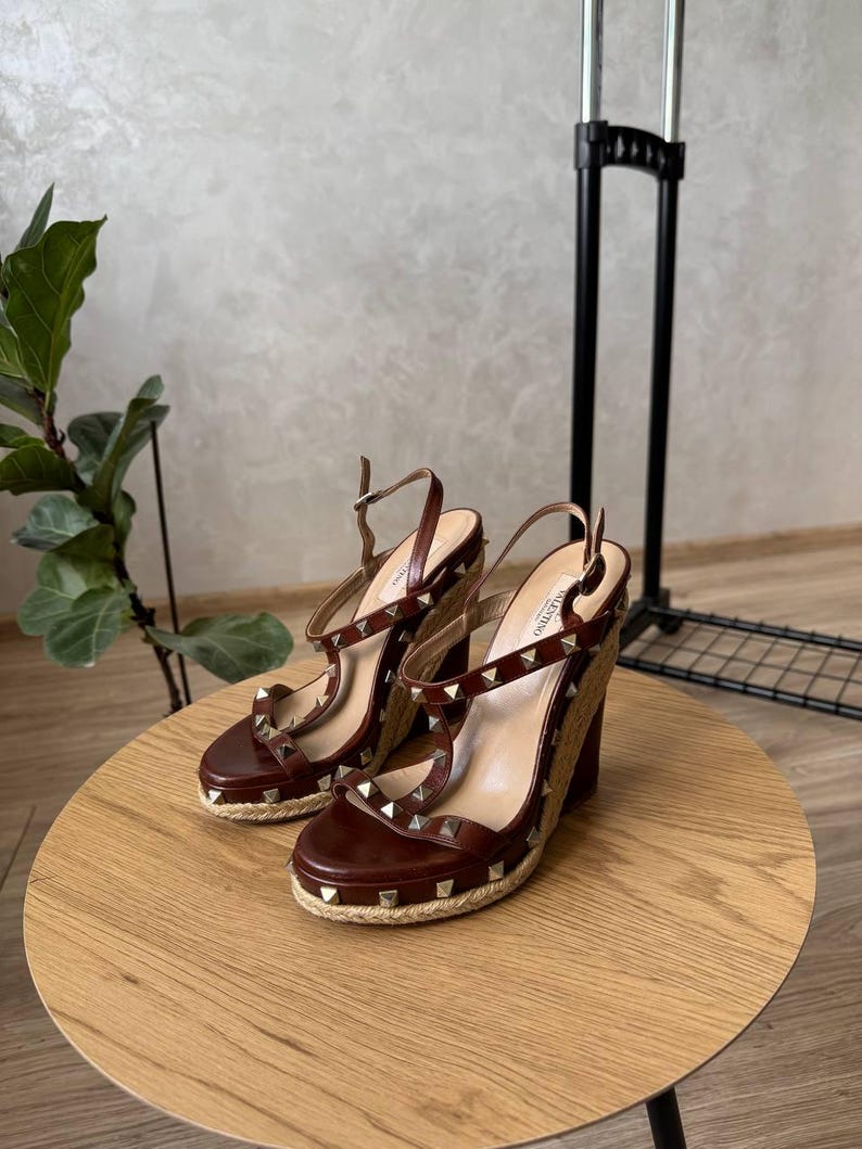 May include: A pair of brown leather wedge sandals featuring a jute-wrapped platform sole, gold-tone pyramid studs along the straps, and an adjustable ankle strap with a metal buckle. The interior footbed is branded with the Valentino logo.