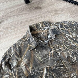 May include: A button-down shirt with a camouflage pattern in shades of brown, green, and tan. The shirt has two chest pockets and a collar. The RealTree Max logo is visible on one of the pockets.