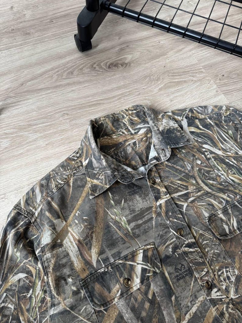 May include: A button-down shirt with a camouflage pattern in shades of brown, green, and tan. The shirt has two chest pockets and a collar. The RealTree Max logo is visible on one of the pockets.