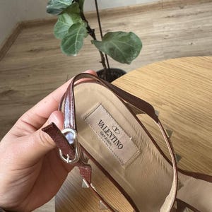 May include: A brown leather sandal featuring a slingback strap with a silver-tone metal buckle. The insole displays a beige fabric label with the text VALENTINO GARAVANI. The strap is adorned with small, evenly spaced silver-tone pyramid studs.