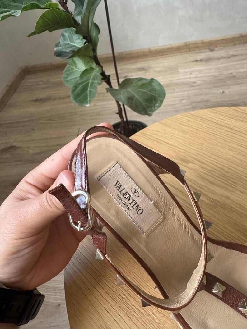 May include: A brown leather sandal featuring a slingback strap with a silver-tone metal buckle. The insole displays a beige fabric label with the text VALENTINO GARAVANI. The strap is adorned with small, evenly spaced silver-tone pyramid studs.