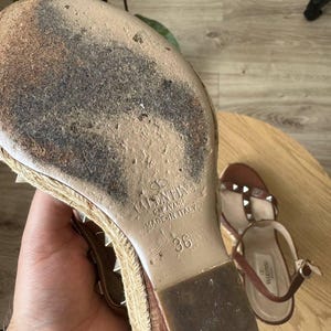 May include: A tan wedge sandal with a jute-wrapped sole and metal studs. The sole is worn and features the embossed text Valentino Garavani Made in Italy and the size 36. A second matching sandal is visible in the background with a brown strap.